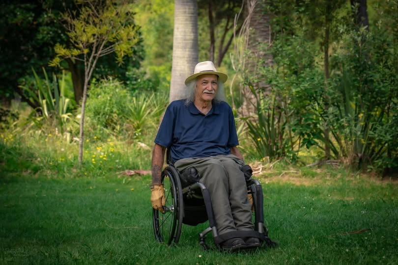 David, smiling softly at the camera, sits in his wheelchair on a green grassy lawn with a densely forested backdrop. 